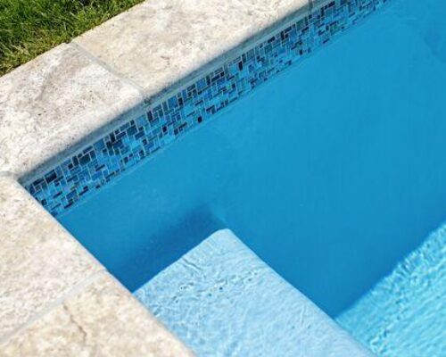 A close-up view of a luxury pool corner with blue water, tiled edges, a submerged step, and grass visible at the top left—perfectly complementing custom spas or outdoor kitchens in your backyard oasis.