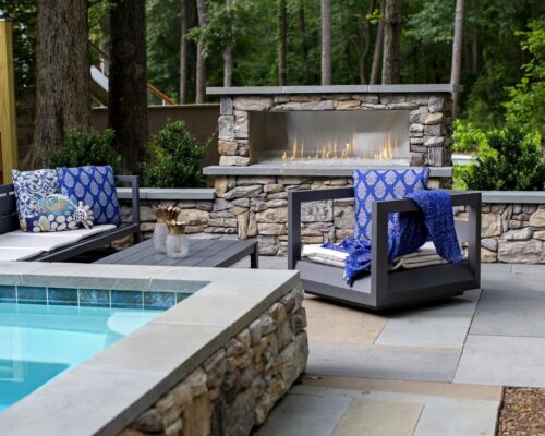 Modern patio featuring a stone fireplace, gray outdoor furniture with blue accents, a coffee table, and an inviting inground pool. Surrounded by lush greenery, it's the perfect space to relax or entertain near custom spas and outdoor kitchens.