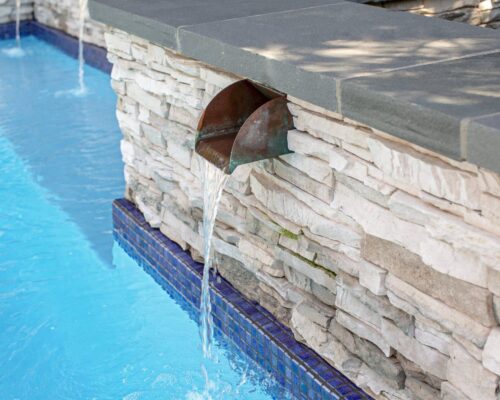 A small waterfall feature built into a stone wall pours water into a clear blue swimming pool lined with blue tiles, creating the perfect setting for luxury pools and custom spas enthusiasts.