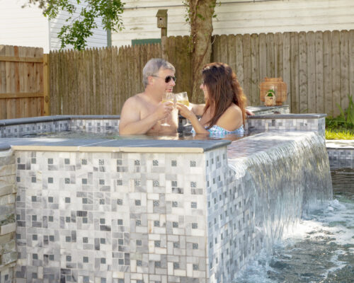 A man and a woman sit in a small tiled inground pool, clinking glasses, with a waterfall edge and a wooden fence in the background.