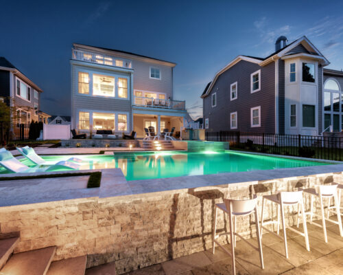 Modern house with large windows and an illuminated swimming pool at dusk, featuring in-pool loungers, patio seating, a row of white barstools at a stone poolside counter, and custom spas for elevated outdoor living.