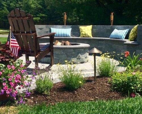 A backyard patio features a stone fire pit, curved stone bench with colorful cushions, an Adirondack chair, flowers, mulch, an American flag, and space perfect for custom spas or outdoor kitchens.