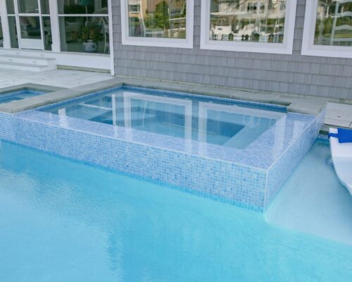 A square hot tub with blue tile is built into a swimming pool next to a house, showcasing custom spas that enhance outdoor living. A pool lounge chair is partially submerged nearby.
