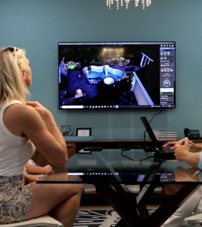 Three people sit at a glass table in a modern office, watching security camera footage of luxurious outdoor kitchens and inground pools displayed on a large wall-mounted monitor.