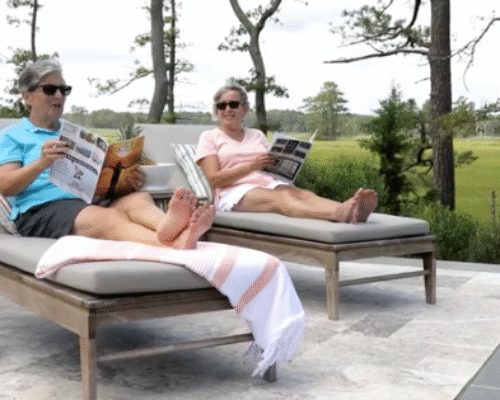 Two people relax on poolside lounge chairs, reading magazines and wearing sunglasses, enjoying the beauty of outdoor living with lush trees and greenery in the background.