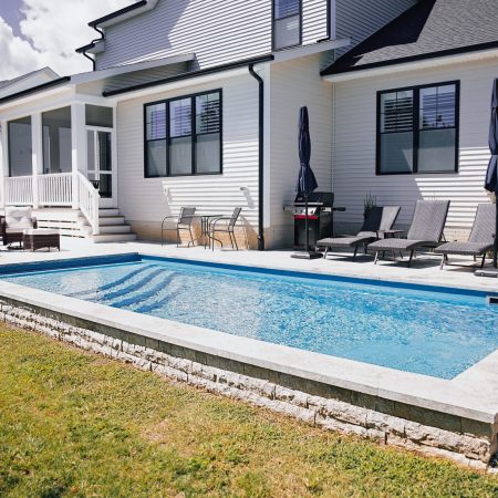 Rectangular inground pool with lounge chairs, umbrellas, and a seating area creates an inviting outdoor living space next to a white house with a screened porch and steps leading to the yard.