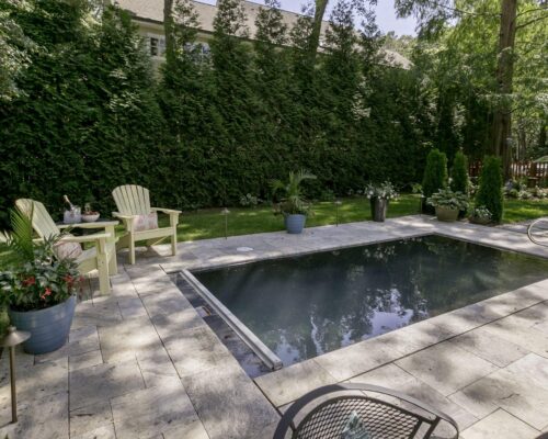 A small rectangular inground pool surrounded by stone tiles, with chairs, potted plants, and trees providing privacy and shade.