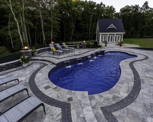Curved luxury pool with water features, surrounded by lounge chairs and a stone patio, plus a small building and trees in the background—perfect for relaxing or entertaining near stylish outdoor kitchens.
