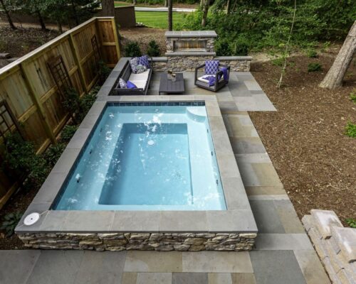 Rectangular hot tub with bubbling water in a fenced backyard patio, adjacent to an outdoor seating area and built-in grill—perfect for enjoying custom spas, surrounded by trees and greenery.