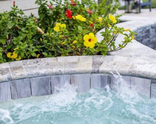 Close-up of a bubbling hot tub with yellow and red hibiscus flowers and green foliage along the stone edge, creating a serene retreat near custom spas or luxurious inground pools.