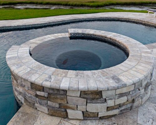 Stone hot tub with raised circular design connected to luxury inground pools; poolside lounge chairs are visible in the background on a stone patio, creating an inviting outdoor living space.