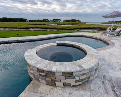 Circular hot tub connected to a fiberglass pool with stone tile decking, lounge chairs, and an umbrella—perfect for outdoor living—overlooking grassy wetlands under a cloudy sky.