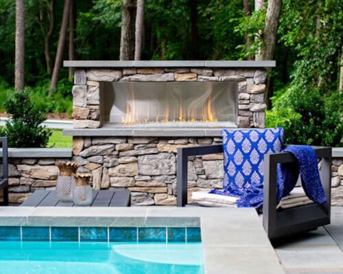 Outdoor seating area with blue cushions and pillows by an inground pool, featuring a stone fireplace in a wooded backyard setting.