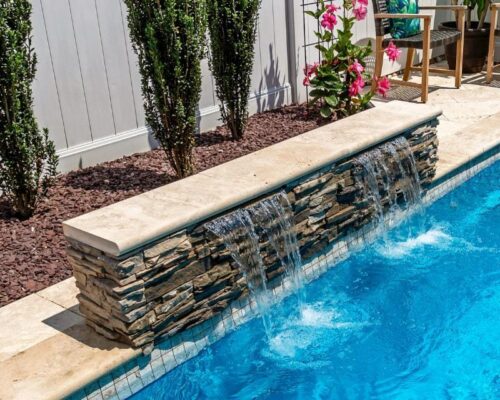 A stone water feature with two waterfalls flows into a blue inground pool. Pink flowers and shrubs are planted along a fence in the background, creating a serene setting for luxury pools and outdoor kitchens.