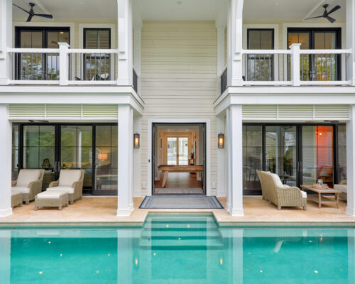 A modern two-story house with balconies overlooks a stylish patio featuring lounge chairs, tables, and a luxury pool—perfect for outdoor living.