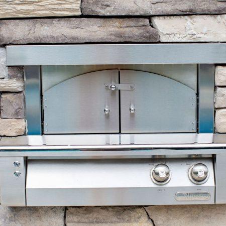 A stainless steel outdoor pizza oven built into a stone wall, perfect for outdoor kitchens, featuring two control knobs and a closed arched door.