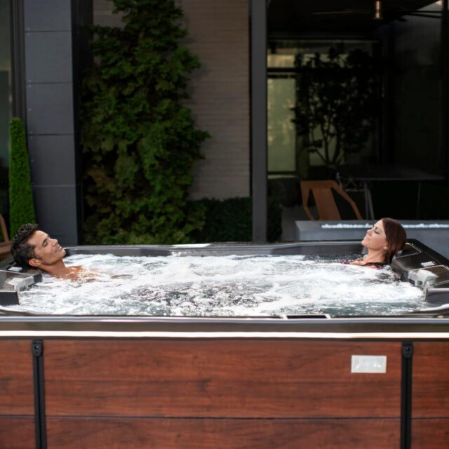 Two people relax in a large outdoor hot tub with bubbling water, set on a patio beside a modern building and lush greenery, showcasing the luxury of custom spas.