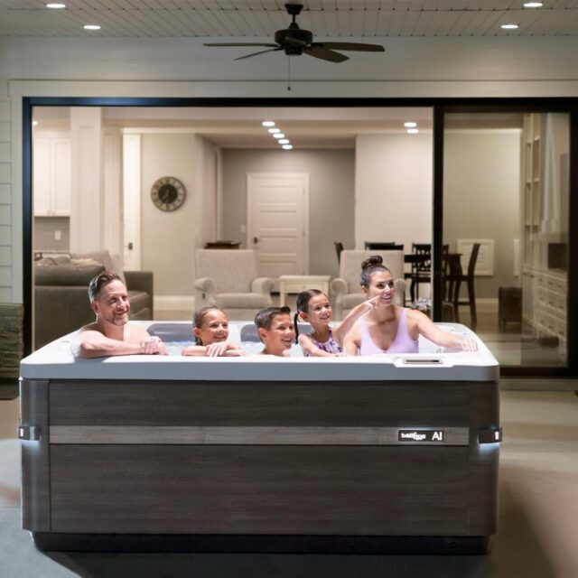 A family of four sits together in a custom spa on a covered patio, with a modern house interior visible through large windows behind them.