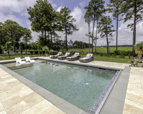 Rectangular backyard inground pool with lounge chairs on a stone patio, surrounded by grass, trees, and a black metal fence—perfect for relaxing or adding custom spas.