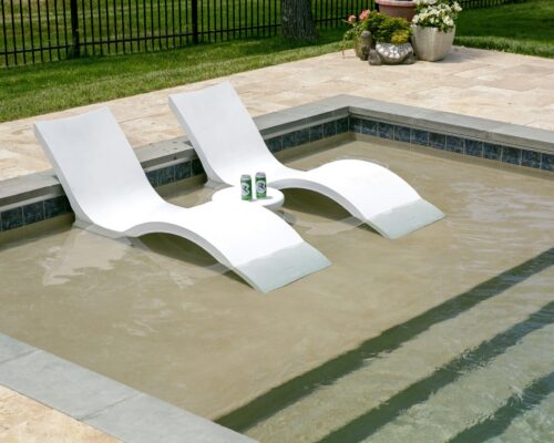 Two white lounge chairs sit in shallow water of a luxury inground pool, with a small round table between them holding two canned drinks; potted plants complete the serene outdoor living scene.