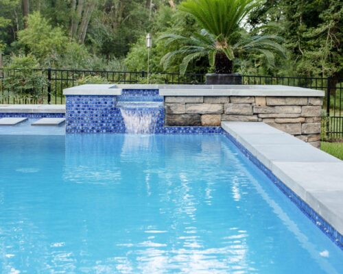 A luxury pool with blue tile accents features a small waterfall, surrounded by stonework and lush landscaping with a potted palm plant—perfect for pairing with outdoor kitchens for the ultimate backyard retreat.