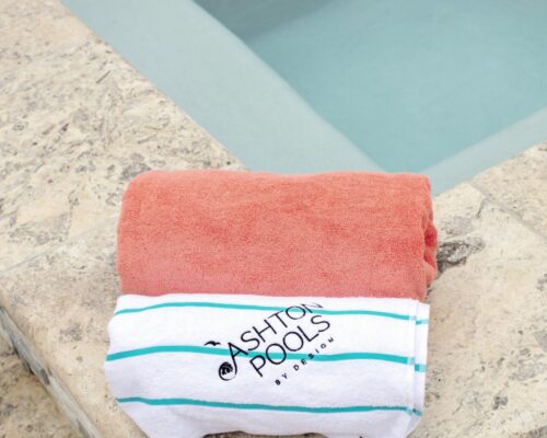 Two folded towels, one coral and one white with blue stripes and Ashton Pools by Design printed on it, rest on the edge of a stone poolside—perfect for enjoying your custom spas or luxurious inground pools in your outdoor living space.