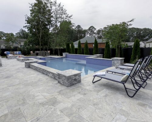 A modern backyard features a rectangular inground pool with stone tile decking, lounge chairs, an outdoor kitchen, and a fire pit seating area surrounded by trees and privacy fencing.