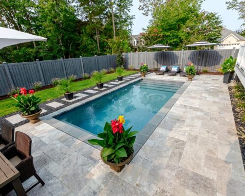Rectangular fiberglass pool with stone patio, lounge chairs, large potted plants, and a wooden fence—perfect for outdoor living amid lush greenery and trees.