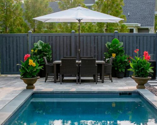A backyard patio with a dining table and chairs under a large white umbrella, surrounded by potted plants, next to a small inground pool.
