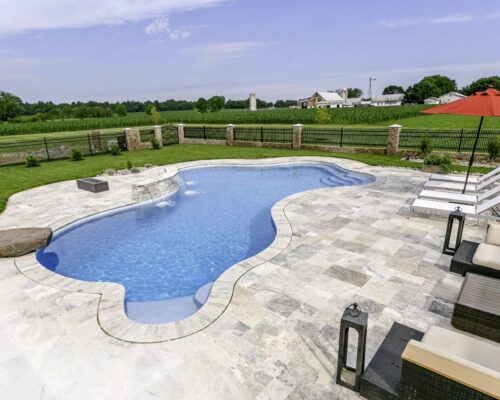 Curved luxury pool with light stone patio, several lounge chairs, a red umbrella, and open green fields in the background.