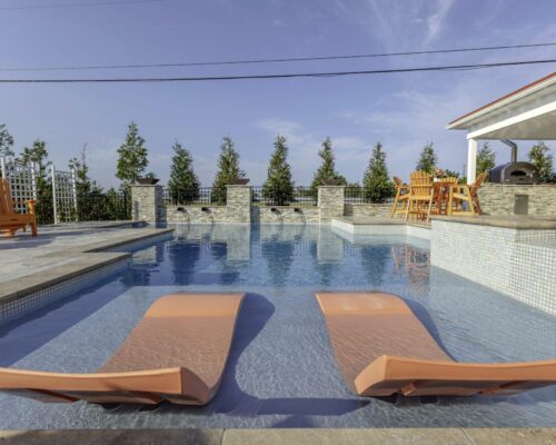Two orange lounge chairs sit in the shallow end of a modern fiberglass pool, with patio furniture and trees in the background under a clear sky, creating the perfect outdoor living space.