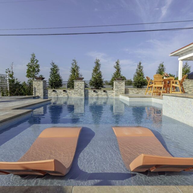 Two orange lounge chairs sit in the shallow end of a modern fiberglass pool, with patio furniture and trees in the background under a clear sky, creating the perfect outdoor living space.