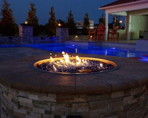 A stone fire pit with flames burns in the foreground near a luxury pool with blue lighting at night, creating the perfect outdoor living ambiance. Trees and patio furniture are visible in the background.