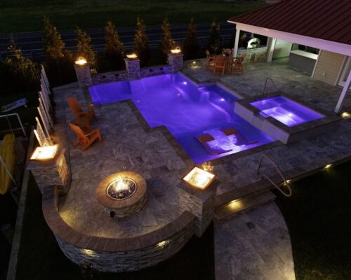 A modern backyard showcases luxury pools, a spa, fire pit, lounge chairs, patio dining set, and a covered outdoor kitchen area at dusk—perfect for stylish outdoor living.