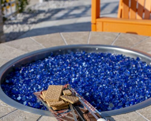 A fire pit with blue glass stones, a tray holding graham crackers and chocolate, and skewers with marshmallows ready for making s'mores sits next to an orange outdoor chair—the perfect setup for relaxing outdoor living.