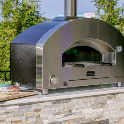 A stainless steel outdoor stone oven sits on a stone countertop, surrounded by pizza ingredients and fresh produce, perfect for enhancing your outdoor living space, with greenery and a road in the background.