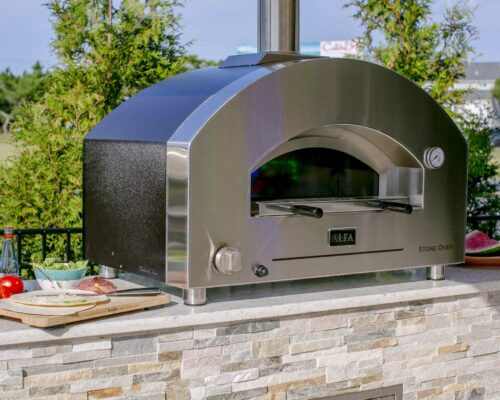 A stainless steel outdoor pizza oven sits on a stone countertop in a stylish outdoor kitchen, surrounded by fresh ingredients and utensils, with greenery, inground pools, and a road visible in the background.
