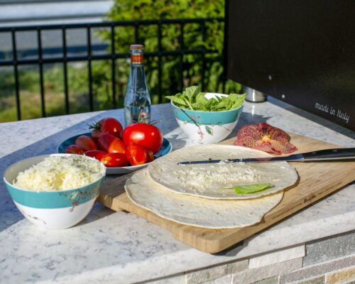 A cutting board with tortillas, shredded cheese, a knife, and a vegetable sits on a counter next to bowls of tomatoes, cheese, greens, and a bottle of sparkling water—perfect for relaxing after enjoying your custom spa or inground pool.