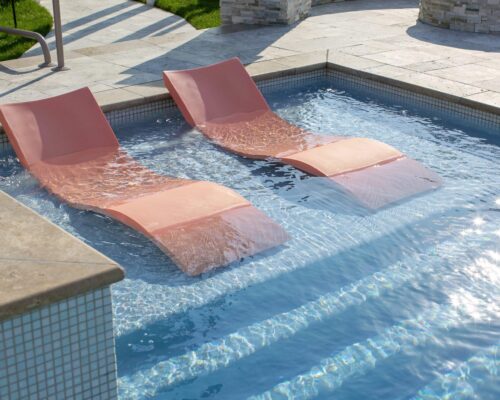 Two pink lounge chairs are partially submerged in the shallow end of a luxury pool, creating the perfect outdoor living vibe on a sunny day.