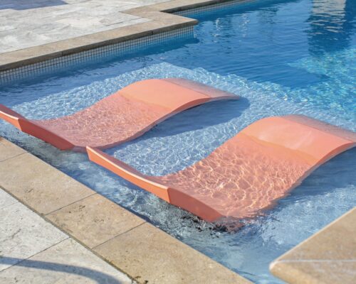Two orange lounge chairs are partially submerged in the shallow end of a luxury pool, surrounded by light-colored stone tiles.