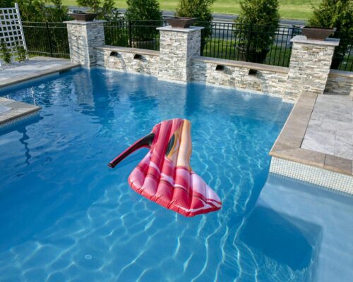 A pink high-heel-shaped pool float drifts in a clear blue inground pool, bordered by stone tiles and a decorative wall with greenery, creating the perfect outdoor living escape.