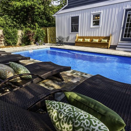 Rectangular luxury pool with wicker lounge chairs, patterned pillows, and a wooden bench set against a white house with glass doors—creating the perfect outdoor living space.