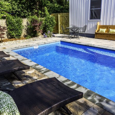 Rectangular backyard swimming pool with lounge chairs, a wooden bench with pillows, and a small patio table set, surrounded by a fenced garden—perfect for those seeking luxury pools or dreaming of custom spas.