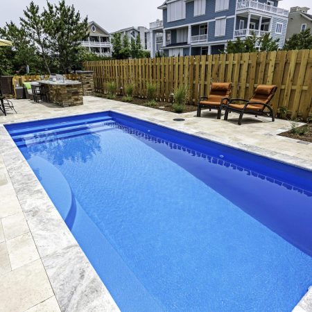 Rectangular backyard fiberglass pool with stone patio, two lounge chairs, a wooden fence, landscaped plants, and houses in the background—perfect for stylish outdoor living.