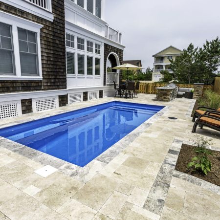 Rectangular luxury pool with stone tile surround, two lounge chairs, outdoor dining area, grill, and wooden fence next to a shingled house—perfect for relaxed outdoor living.