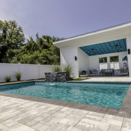 Rectangular inground pool with stone tile decking, a small waterfall feature, and a modern white pool house with blue ceiling and seating area—perfect for outdoor living. White privacy fence and trees create a serene backdrop.
