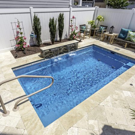 A small rectangular luxury pool with a stone water feature, surrounded by patio chairs, plants, a white privacy fence, and space for outdoor kitchens to enhance your backyard retreat.