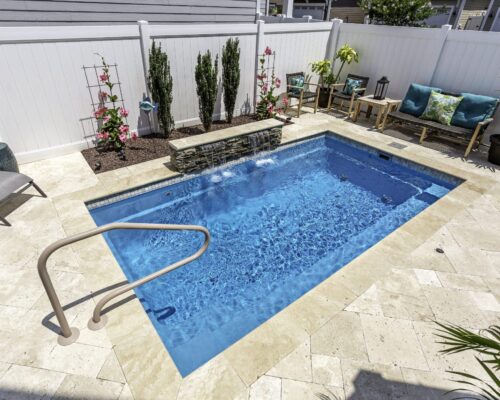 A small rectangular luxury pool with a stone water feature, surrounded by patio chairs, plants, a white privacy fence, and space for outdoor kitchens to enhance your backyard retreat.