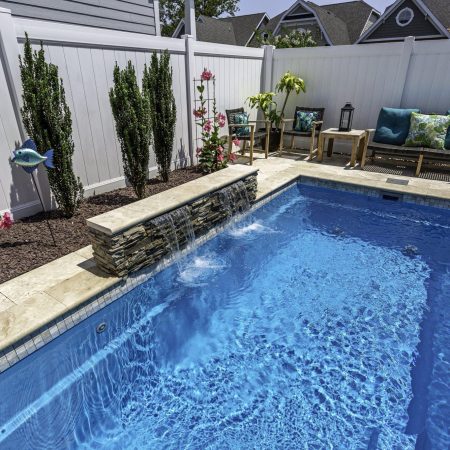 Small backyard luxury pool with a stone waterfall feature, surrounded by plants and patio seating, enclosed by a white privacy fence.