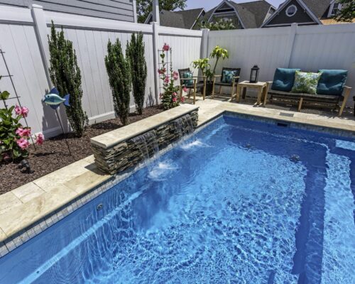 Small backyard luxury pool with a stone waterfall feature, surrounded by plants and patio seating, enclosed by a white privacy fence.